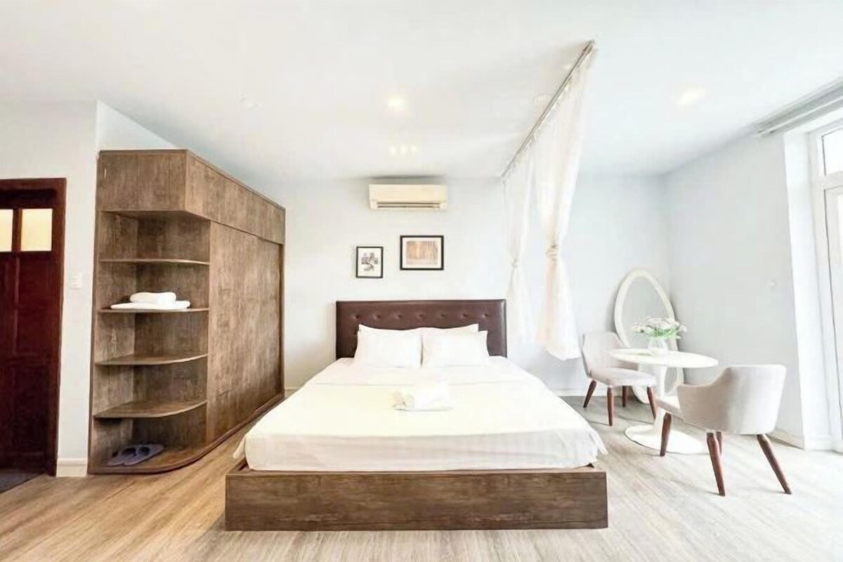 A modern hotel room with a large bed, a wooden wardrobe, a small desk with a chair, and a large window.