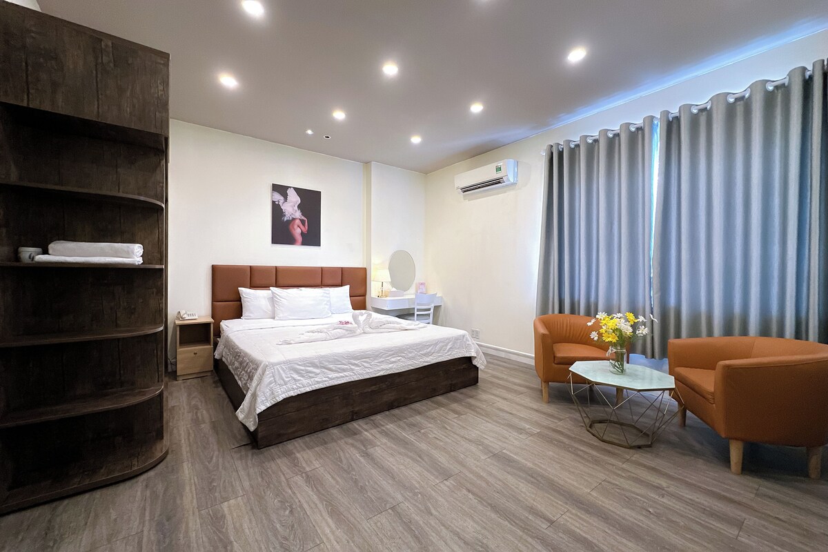 A modern hotel room with a large bed, a wooden wardrobe, a seating area with an orange sofa and a small table, and a large window with curtains.