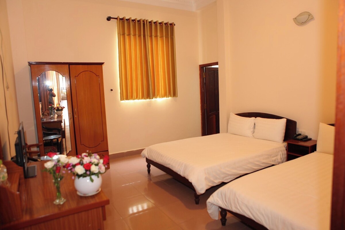 A hotel room with two beds, a wooden wardrobe, a desk with a vase of flowers, and a window with curtains.