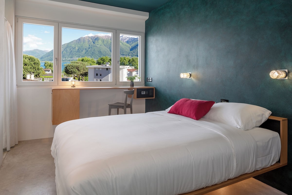 Un dormitorio con cama, escritorio y silla, ventana con vistas a montañas y pared de color aguamarina.