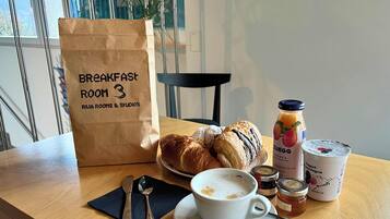 Ein Frühstückstisch mit Croissants, einer Kaffeetasse, einem Honigglas, einer Saftflasche und einer Papiertüte mit der Aufschrift 'Frühstückszimmer 3'.