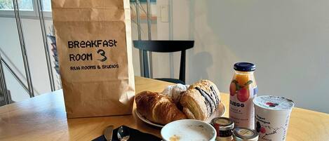 A breakfast table with croissants, a coffee cup, a jar of honey, a bottle of juice, and a paper bag labeled "Breakfast Room 3".