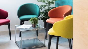 Una habitación con cuatro sillas en rojo, verde azulado, naranja y amarillo, una mesa de vidrio con una planta y una pared azul.