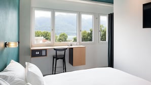 A bedroom with a bed, a desk, and a chair. There are three windows providing a view of mountains and trees. - Riva Rooms & Studios (Minusio)