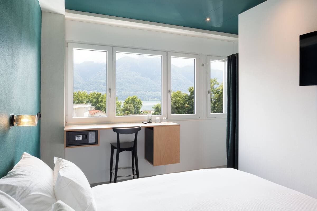 Un dormitorio con cama, escritorio y silla. Se ven tres ventanas que dan a vistas de montañas y árboles.