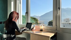 A person working at a desk by a window with a view of mountains and a lake. - Riva Rooms & Studios (Minusio)