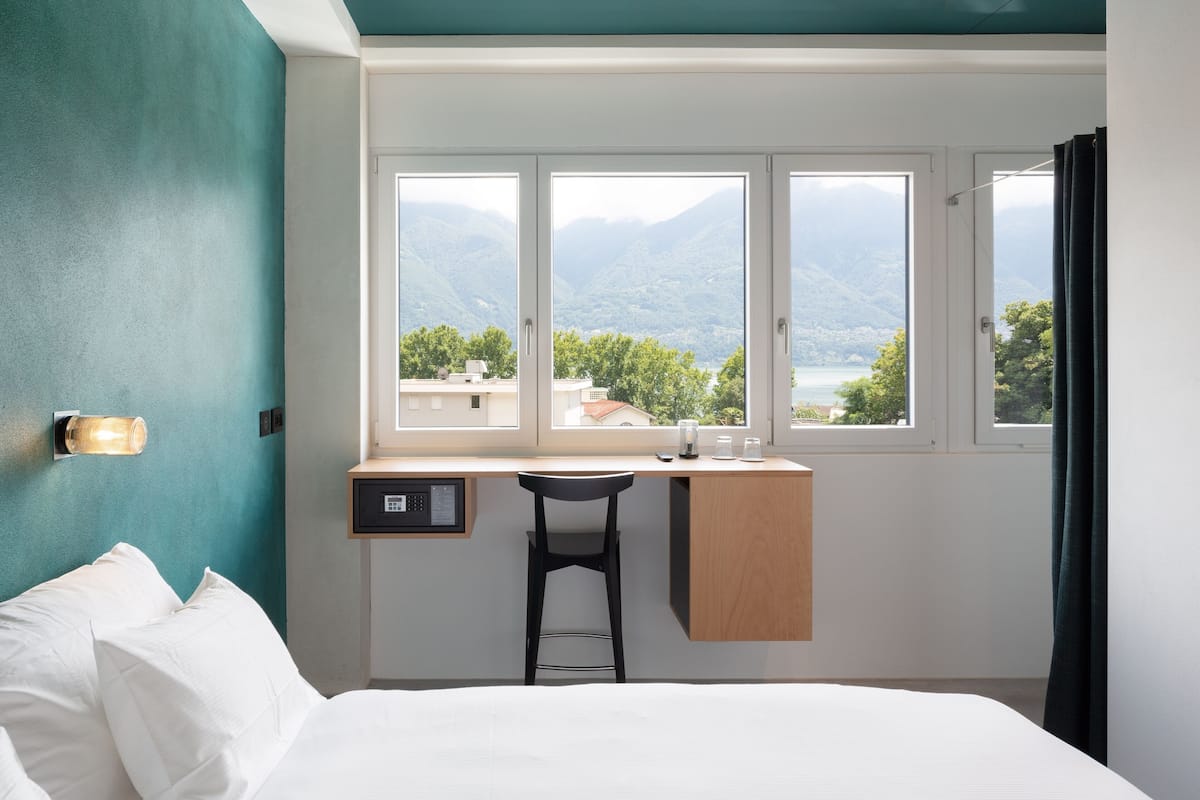 Un dormitorio con cama, escritorio y silla. Se ve una ventana con vistas a montañas y un lago.