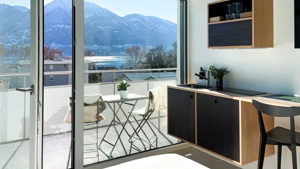 A modern kitchen with a view of mountains, a dining area with a table and chairs, and a small balcony with a plant. - Riva Rooms & Studios (Minusio)