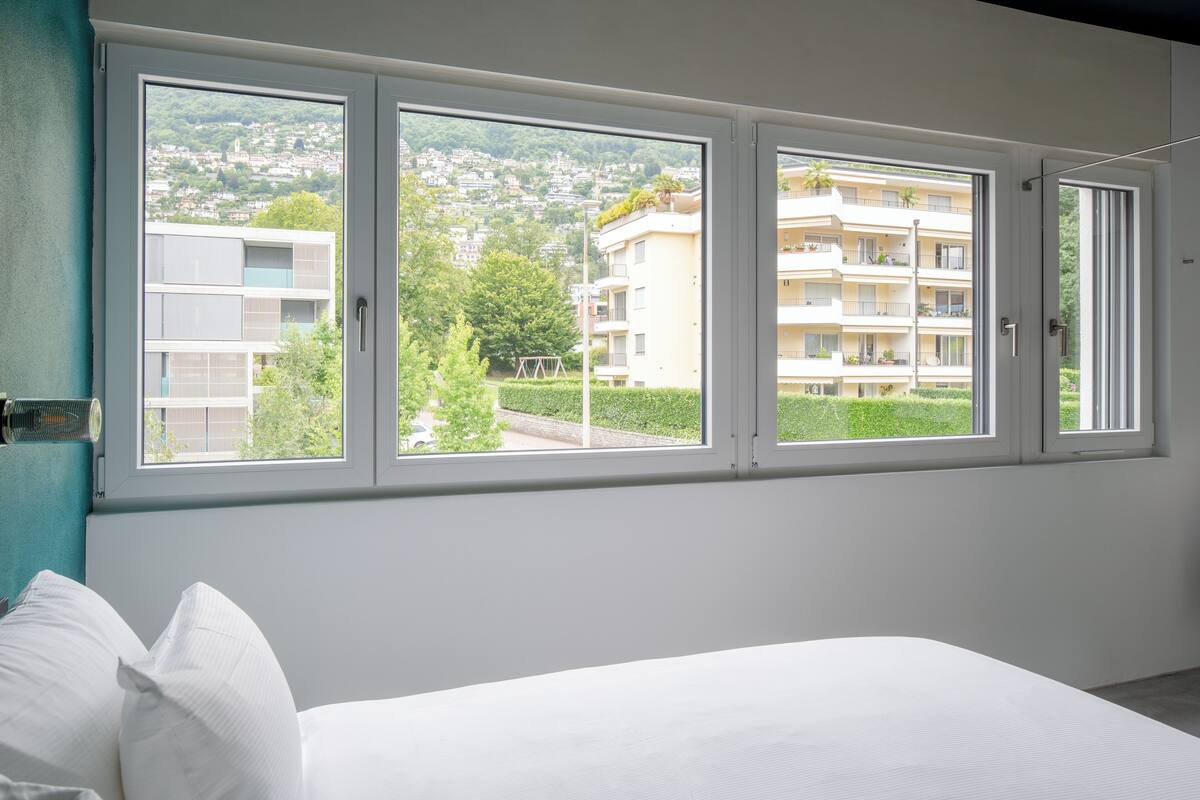 Un dormitorio con un ventanal grande, ropa de cama blanca y vistas a una zona residencial.