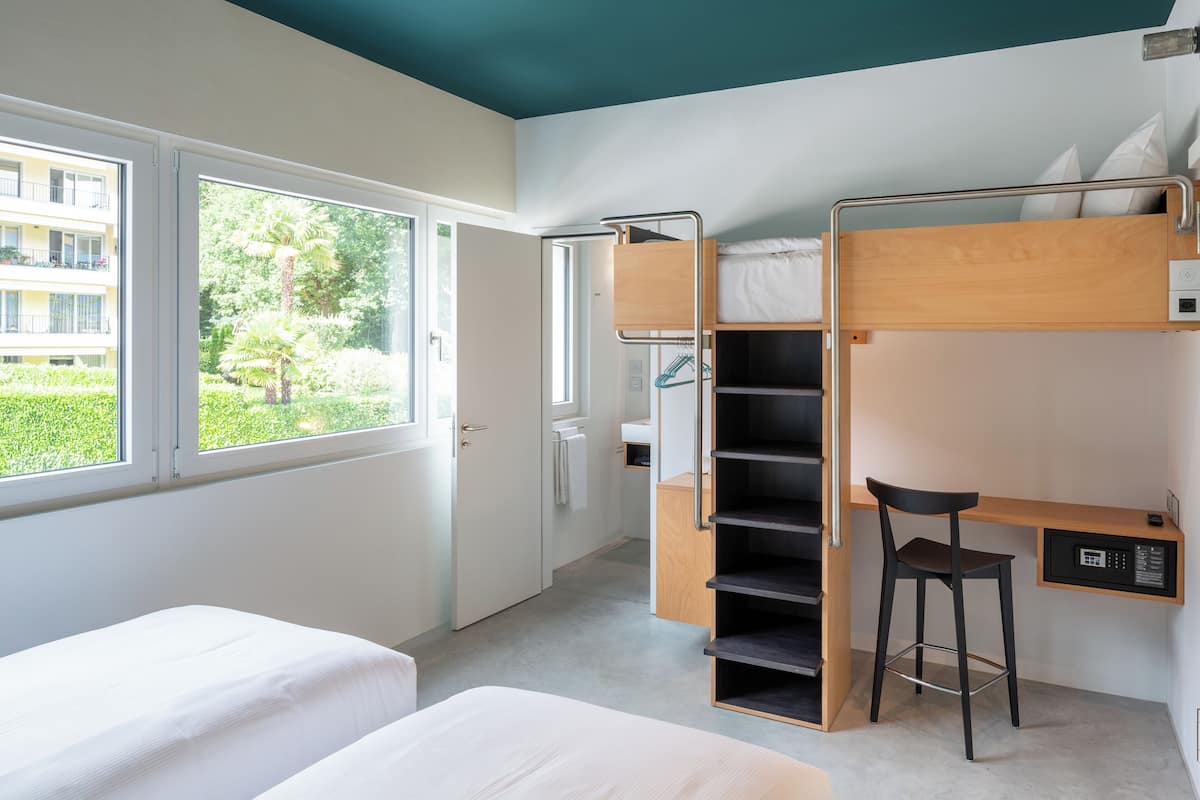 Una habitación de hotel moderna con una cama litera, un escritorio con silla y un amplio ventanal que ofrece vistas al verdor.