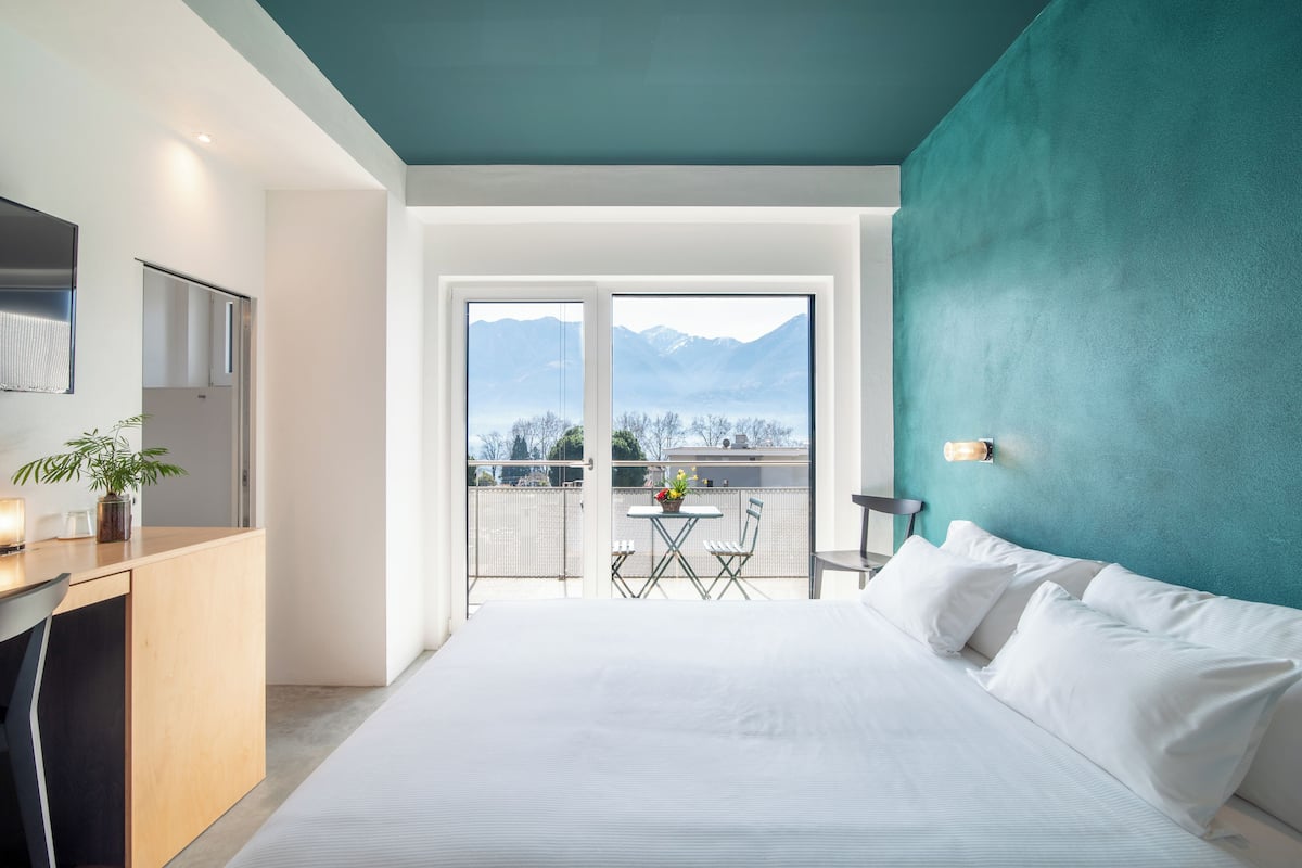 Un hotel moderno con una cama grande, un escritorio con silla, una pequeña mesa con sillas y vistas a montañas a través de las puertas del balcón.