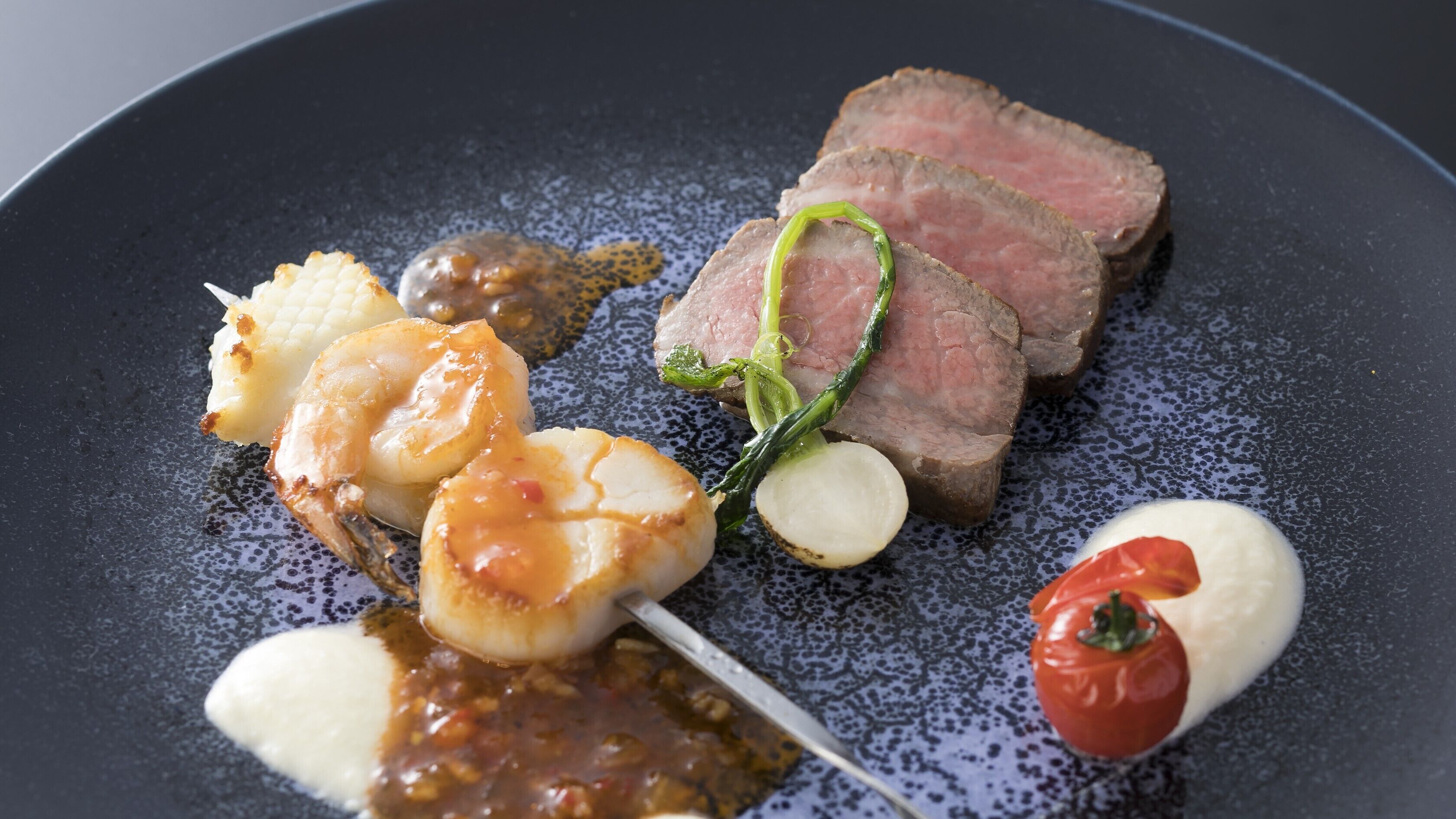 Une assiette contenant des tranches de viande, desSaint-Jacques, une tomate et un petit légume.