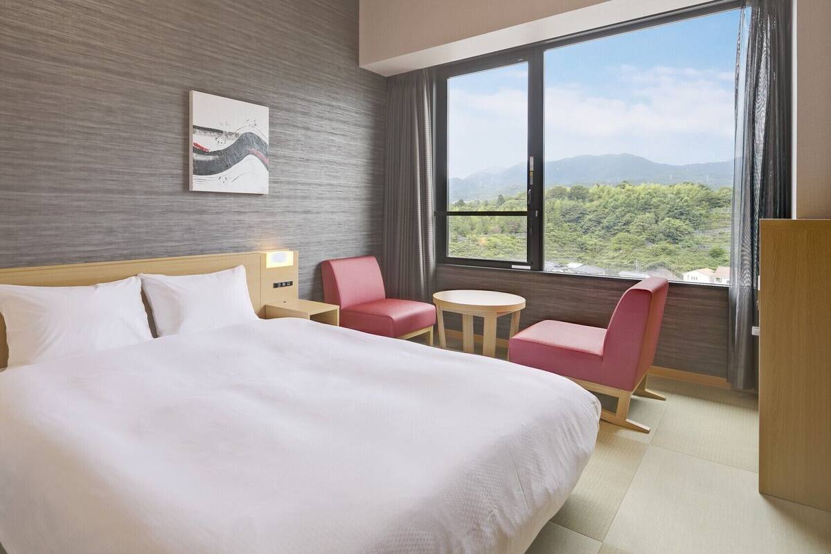 A hotel room with a large bed, a desk, a chair, a small table, and a view of greenery and mountains.
