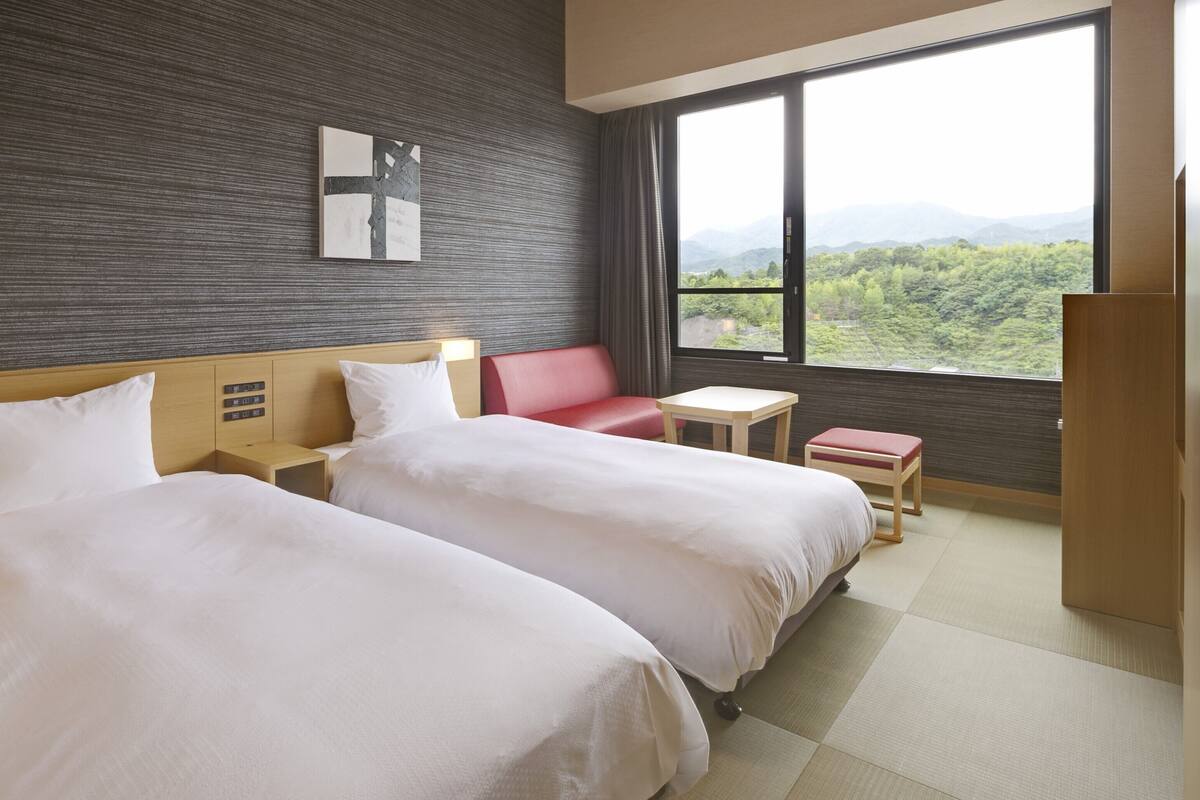 A hotel room with two beds, a small table, a red chair, and a large window offering a view of the mountains.