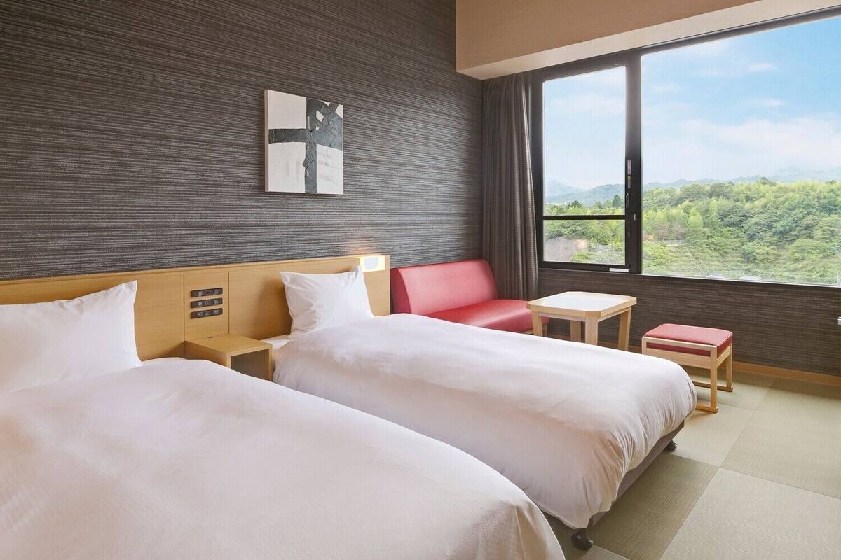 A hotel room with two beds, a small table, a red chair, and a view of greenery through the window.