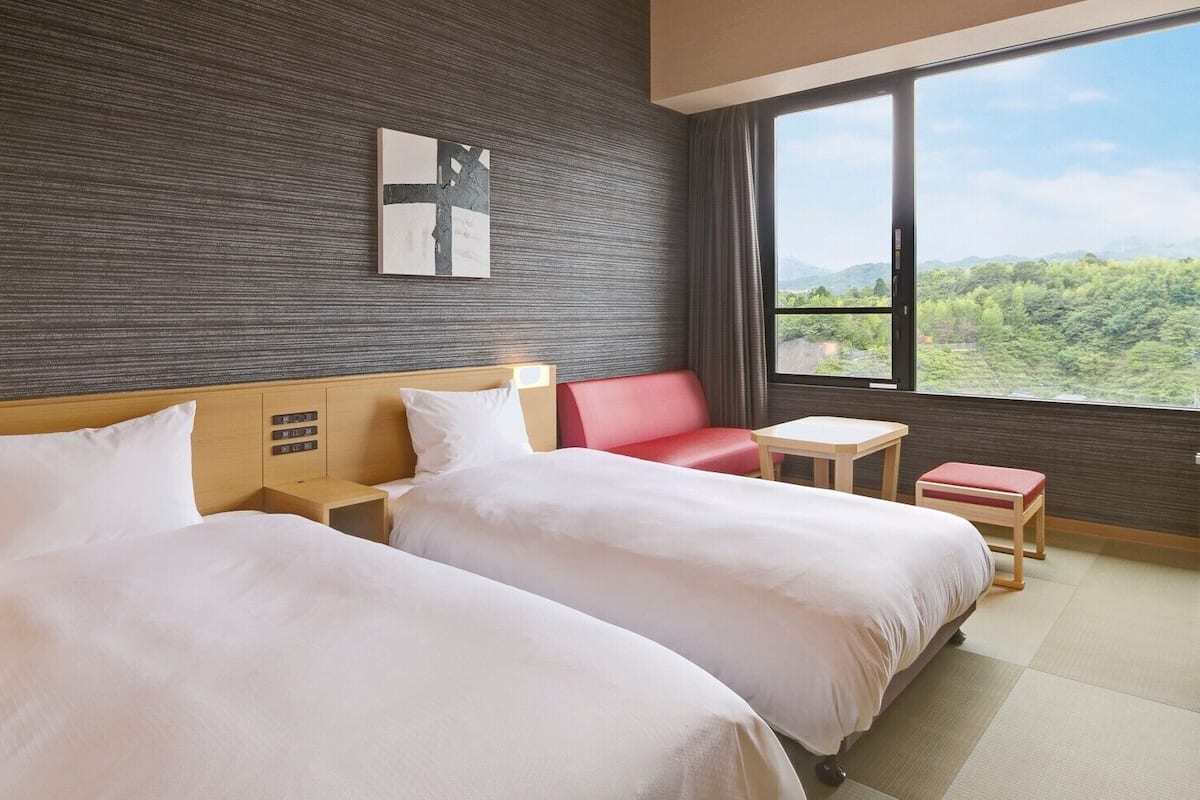 Habitación de hotel con dos camas, una mesita, una silla roja y vistas al verdor por la ventana.