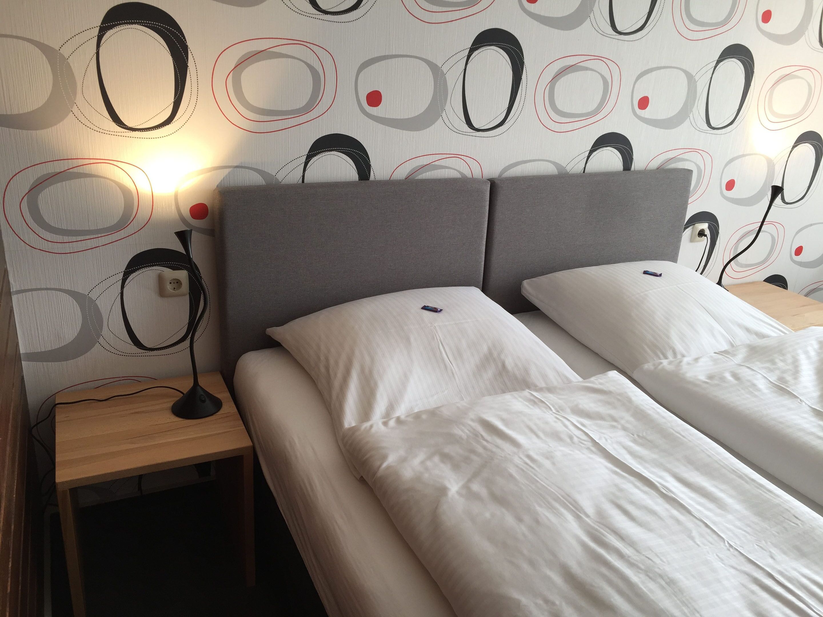 A double bed with white linens, a grey headboard, and a bedside table with a lamp.