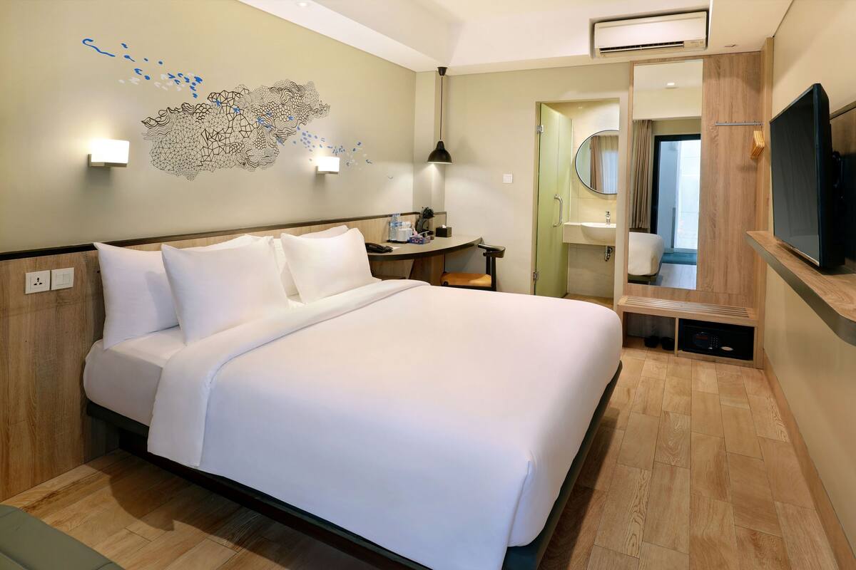 A hotel room with a large bed, a desk with a lamp, a TV, and a bathroom visible through an open door.