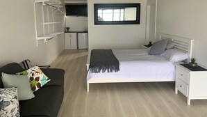 Quarto com cama, mesa de cabeceira e um pequeno criado-mudo branco. Há uma janela com persianas, um aparelho de ar condicionado branco e uma área de kitchenette ao fundo.