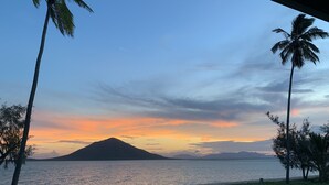 Vista do pôr do sol de um espaço coberto, com palmeiras e uma montanha ao longe.