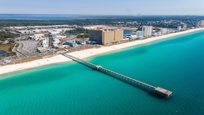 Vue aérienne d’une zone côtière avec une longue jetée s’étendant dans des eaux turquoise, une plage de sable et plusieurs immeubles de grande hauteur en arrière-plan.