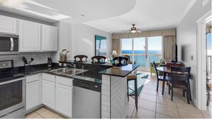 A modern kitchen with white cabinetry, a central island, and a dining area with a view of the ocean. - Calypso 1005E - Gulf-Front Luxury, Beach Svc Included, Pier Park, Sleeps 4 (Panama City Beach)