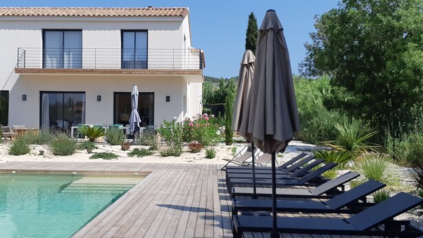 A modern two-story building with a swimming pool, sun loungers, and umbrellas.