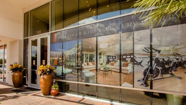 A modern building with large glass windows reflecting an interior with a fitness area, sofas, and a coffee table. - Spacious Furnished Apartment by NRG Stadium (Houston)