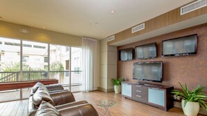 A modern living room with a leather sofa, a glass coffee table, a TV mounted on a wooden wall, and a balcony with a railing. - Spacious Furnished Apartment by NRG Stadium (Houston)