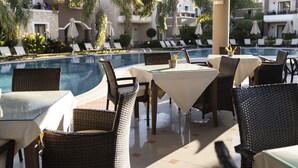 Un área de comedor junto a la piscina con sillas y mesas de mimbre, con vistas a la piscina y a los edificios del hotel.