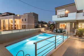 A swimming pool with a metal railing, surrounded by buildings with balconies and outdoor seating. - Okeanis 2 (Chania)