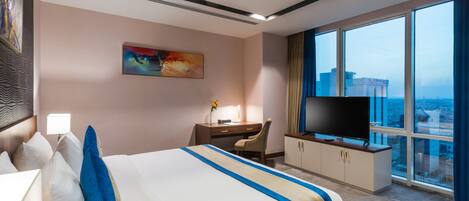 A hotel room with a large bed, a desk with a chair, a television, and a large window offering a city view.