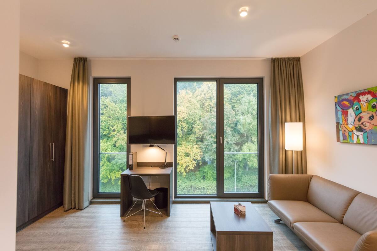 Una habitación moderna con escritorio, silla, sofá y ventanas amplias que dan a zonas verdes.