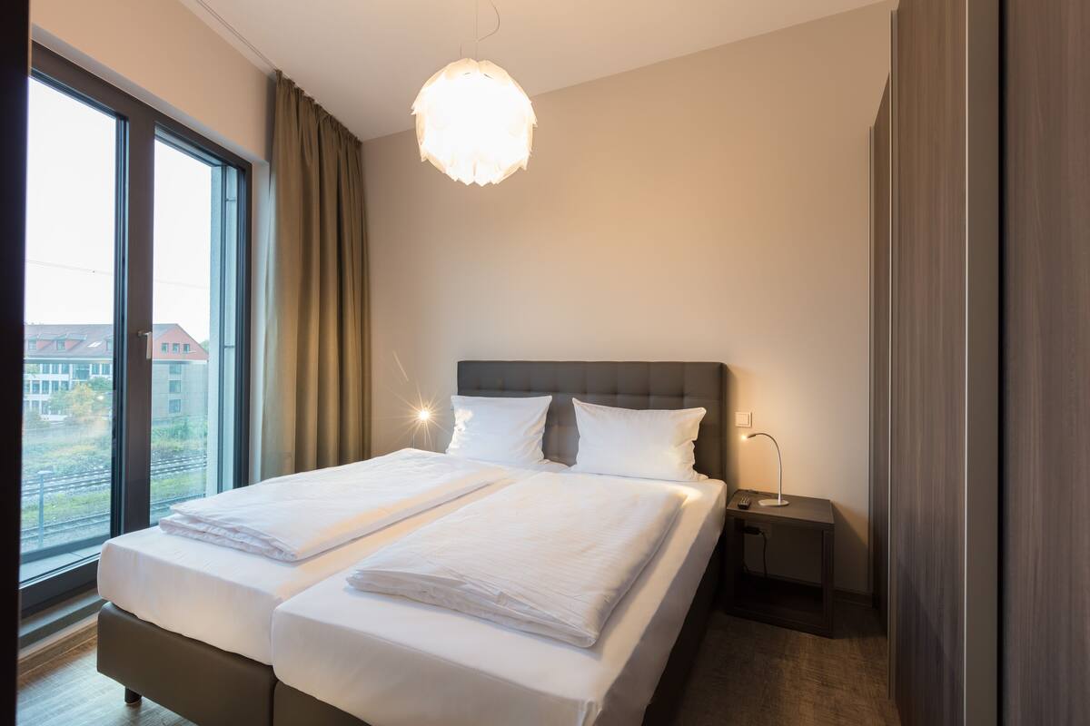 Habitación de hotel con una cama grande, una mesita de noche, una lámpara y vistas al exterior.