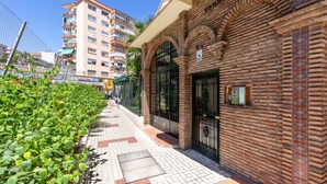 Un edificio de ladrillo con una entrada protegida por una reja, un sendero pavimentado y una fila de plantas verdes.