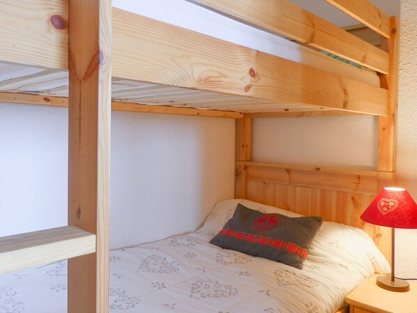 A bunk bed with a wooden headboard, a single bed with a floral-patterned bedspread, a red and black pillow, and a bedside table with a red lamp.