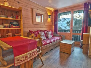 A cozy cabin interior with a wooden interior, a dining table with a red tablecloth, a sofa with heart-patterned cushions, and a view of a snowy landscape through the window.