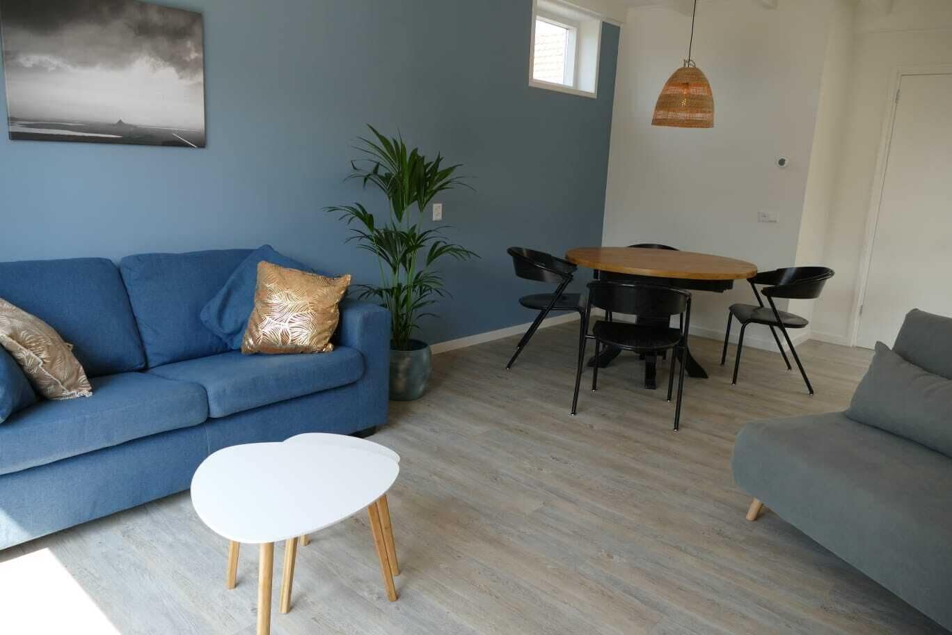 Un salon avec un canapé bleu, une table basse blanche, une table à manger avec des chaises, un luminaire suspendu et une plante en pot.