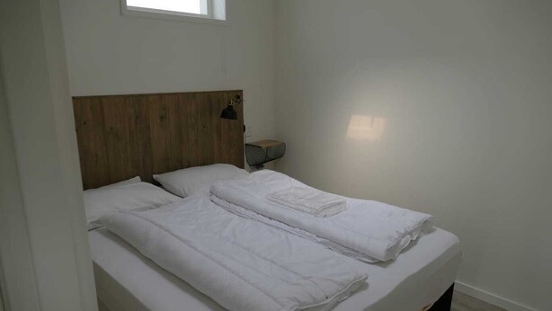A small, clean room with a single bed, a wooden headboard, a bedside table with a lamp, and a window providing natural light.