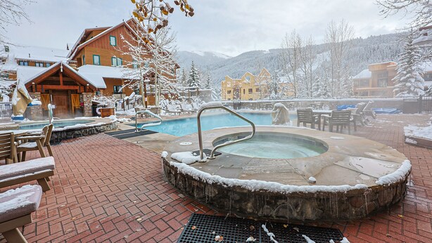 A hot tub area with a pool, wooden benches, and a stone hot tub surrounded by snow-covered trees and buildings. - River Run Village Walk To Gondola Pool Hot Tubs Restaurants and Shops DELUXE (Keystone)