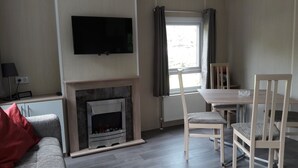 Uma sala de estar com lareira, televisão na parede, mesa de jantar com cadeiras e janela com cortinas.
