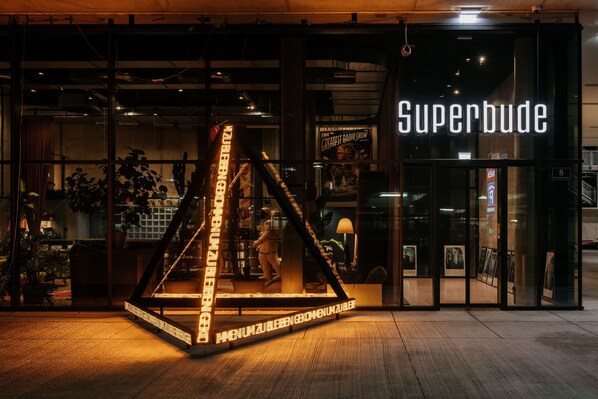 La entrada de un edificio con un letrero de neón que dice 'Superbude' y una estructura triangular con texto iluminado.