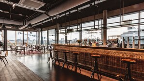 Zona de bar moderna con techos altos, conductos expuestos y grandes ventanales con vistas a la ciudad.