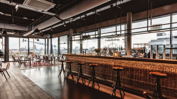 A modern bar area with high ceilings, exposed ductwork, and large windows offering a city view. - Superbude Wien Prater (Vienna)