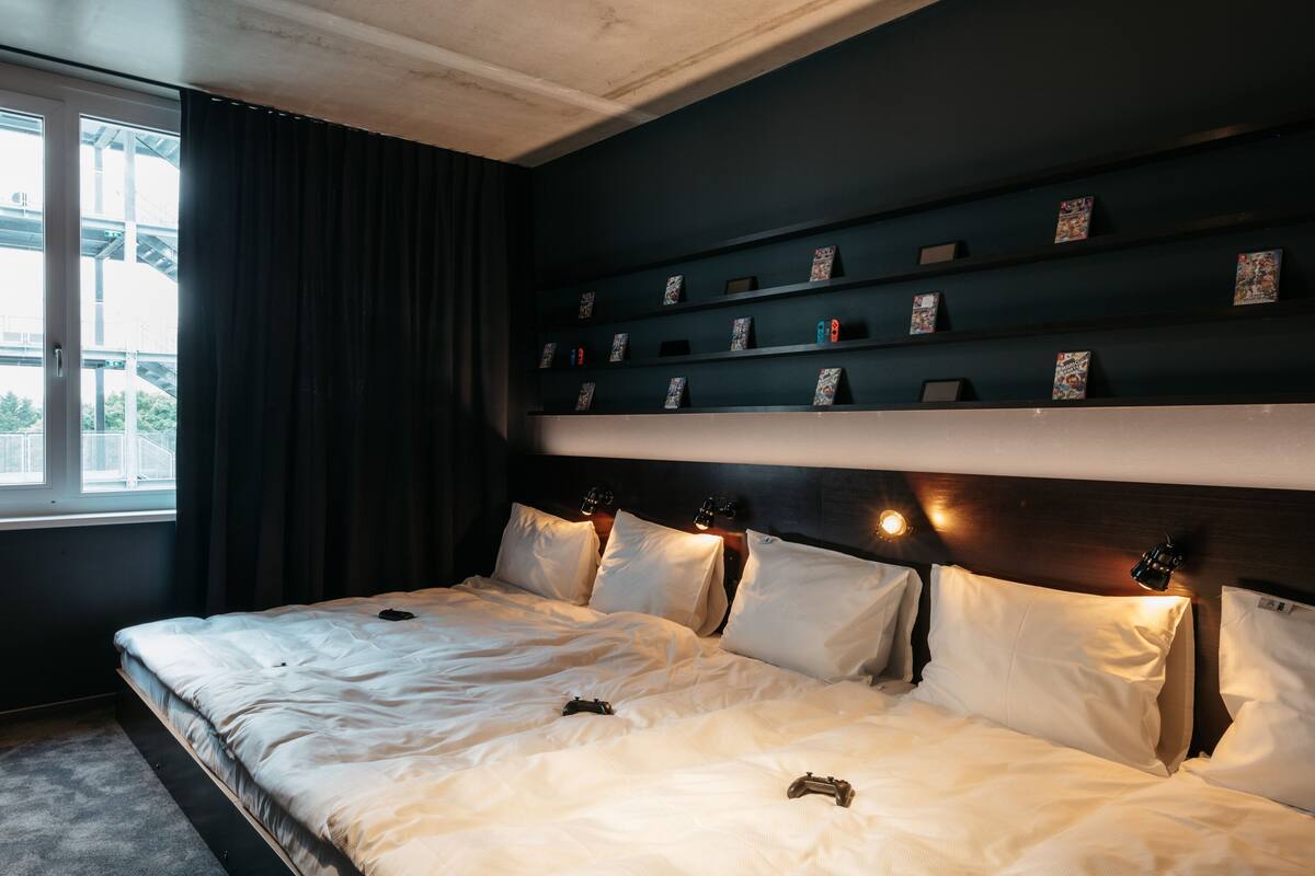 A hotel room with a large bed, a headboard with built-in shelves displaying items, a window with curtains, and a view of an outdoor structure.