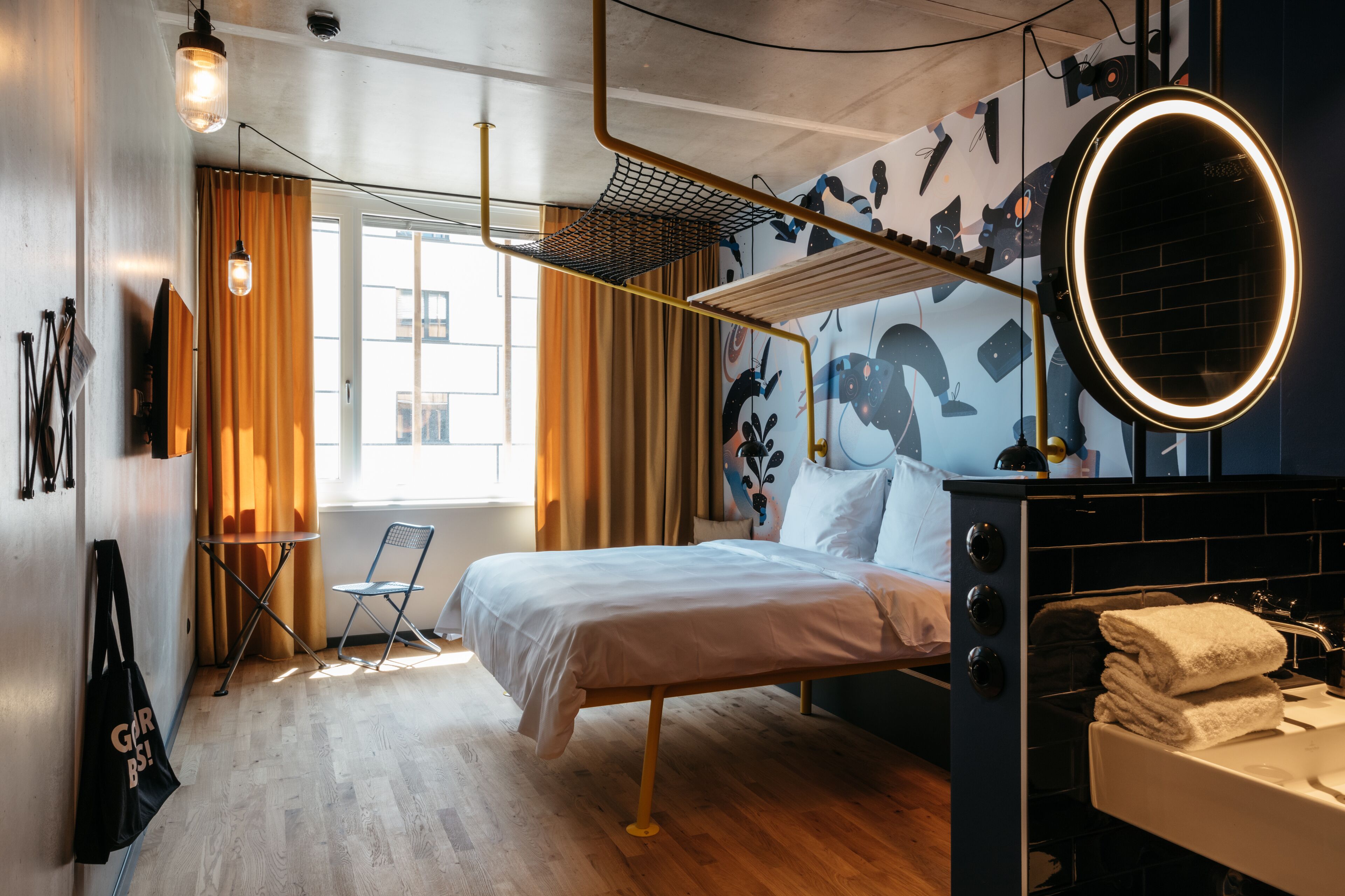 A modern hotel room with a bed, a small table, a chair, a bathroom with a mirror, and a window with curtains.