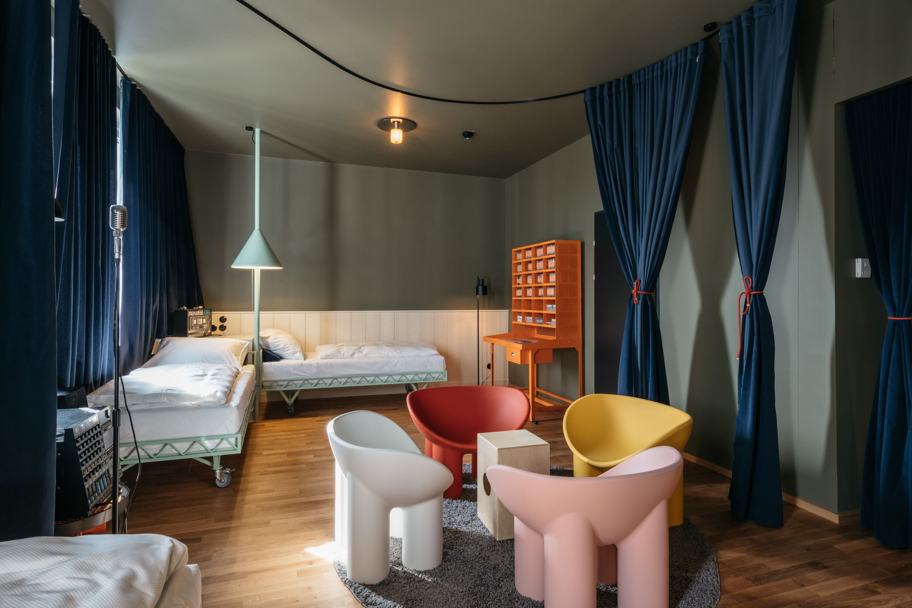 A modern hotel room with a curved ceiling, wooden flooring, two beds, a red chair, a yellow chair, a white chair, a small table, and a lamp.