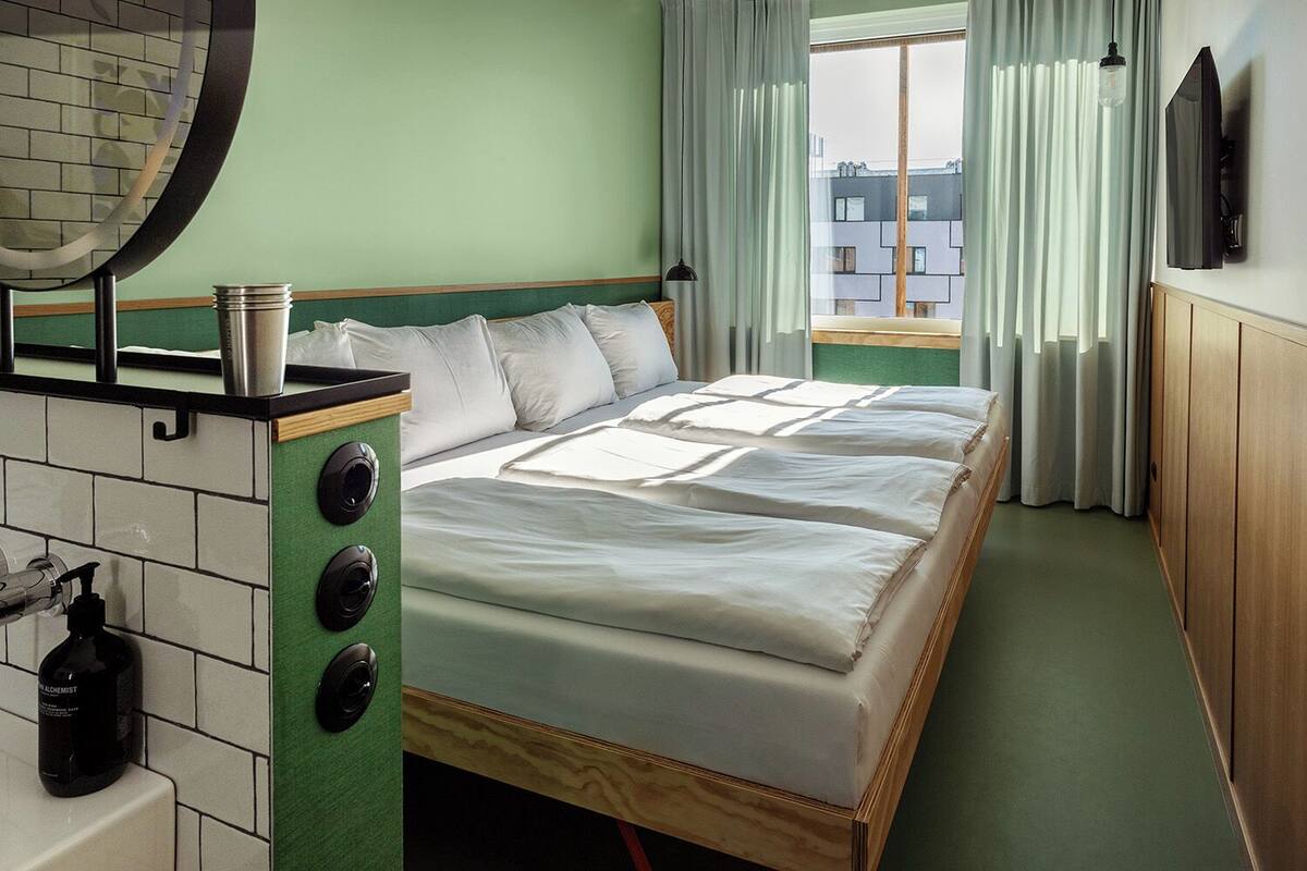 A hotel room with a bed, a small table with a kettle, a mirror, and a window with curtains.