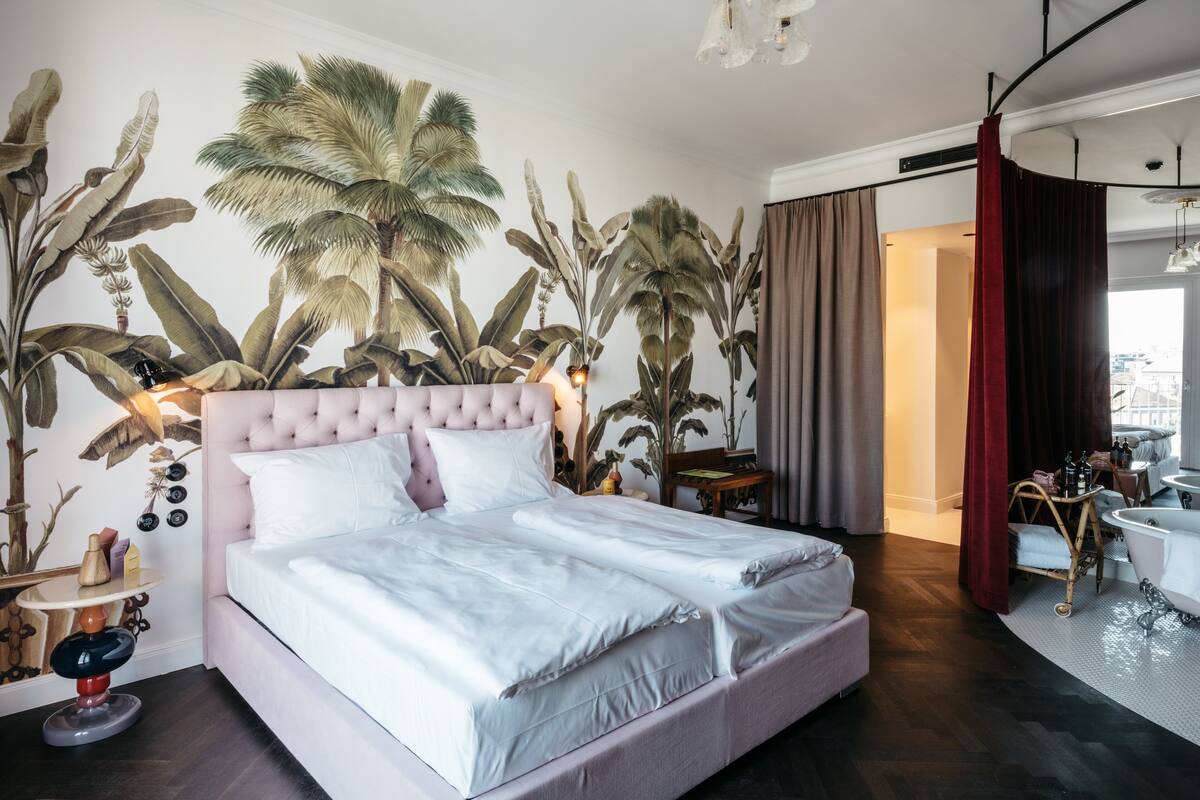 A bedroom with a large bed, a nightstand, a chair, a bathroom with a bathtub, and a large plant wall.