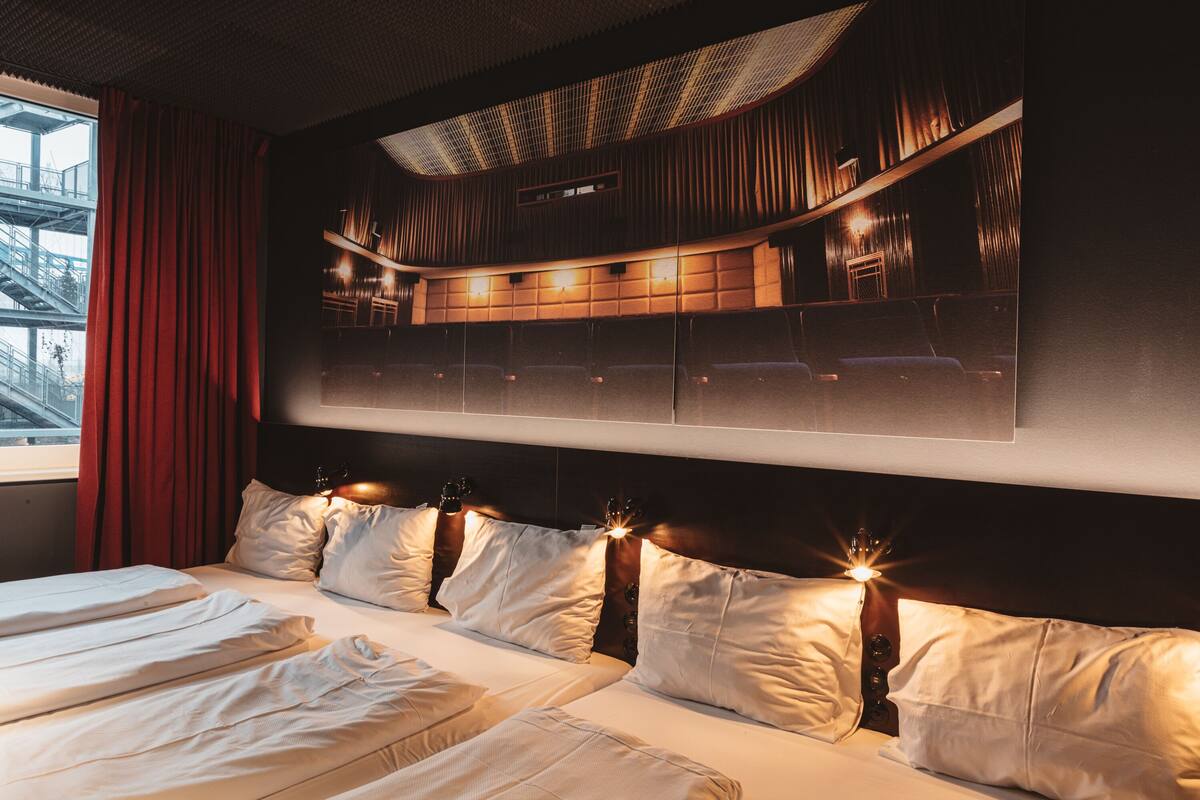A hotel room with a large bed, a headboard with built-in speakers, and a view of a cityscape through the window.